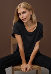 Blondýna v černé košili a kalhotách sedí na dřevěné židli před hnědým pozadím, naklánějící se dopředu se sepnutýma rukama.