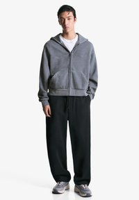 Šedá mikina na zips s prednými vreckami, nosená cez bielu košeľu, v kombinácii s čiernymi teplákmi a šedými športovými topánkami. Hladká textura, uvoľnený strih.