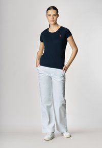 Una donna sta in piedi con sicurezza, indossando una t-shirt blu navy con un piccolo logo rosso, pantaloni bianchi a gamba larga e sneaker bianche, con le mani in tasca.