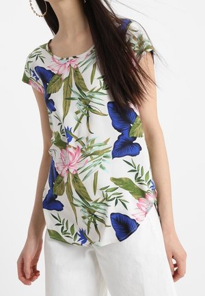 Kobieta nosząca biały krótki top z zielonymi liśćmi, różowymi kwiatami i dużymi niebieskimi motylami, w połączeniu z białymi spodniami.