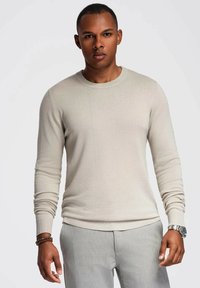 Maglione beige chiaro con scollatura circolare, maniche lunghe e design aderente, abbinato a pantaloni grigi chiari e un orologio scuro.