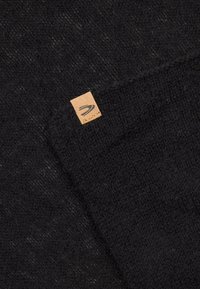 Schwarzer gestrickter Stoff mit einer weichen Textur, versehen mit einem hellbraunen Lederetikett mit einem Logo. Das Design ist einfach und minimalistich.