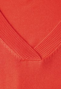V-Ausschnitt-Pullover in lebhaftem Koralle, mit geripptem Kragen und feiner Strickstruktur, nahtloser Verarbeitung entlang der Kanten.