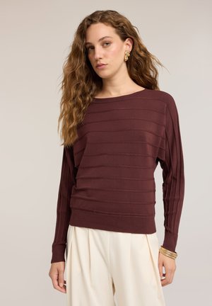 Junge Frau mit lockigem Haar, die einen bordeauxroten gerippten Pullover, cremefarbene weite Hosen, goldene Ohrringe und Armbänder vor einem schlichten Hintergrund trägt.