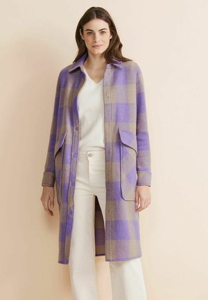 Long, button-up outerwear in a purple and beige plaid pattern. Soft fabric with two front pockets and a collar. White top and light pants.