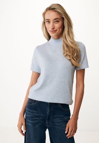 Žena s dlouhými blond vlasy, oblečená v svetru s krátkým rukávem světle modré barvy a tmavě modrých džínách, stojící na bílém pozadí.