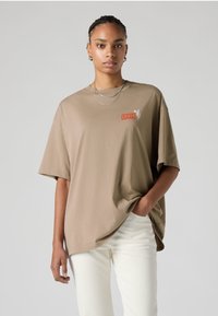 Beige oversized cotton t-shirt with short sleeves and a red Levi's logo featuring a cactus graphic on the left chest. White pants.