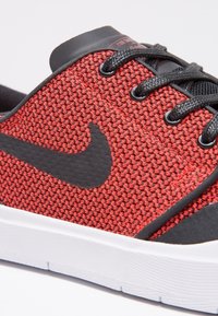 Baskets en tissu rouge tissé avec des accents noirs, présentant un grand logo swoosh noir, des lacets ronds et une semelle en caoutchouc blanche.