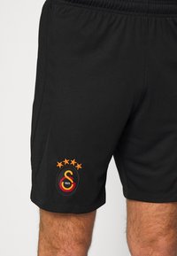 Short de sport noir avec une ceinture élastique, arborant le logo de Galatasaray avec quatre étoiles orange sur la jambe gauche.