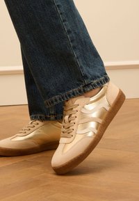 Baskets de sport beiges et dorées avec une tige en suède et métallique, dotées d'une semelle en caoutchouc et d'un laçage. Portées avec un pantalon en denim.