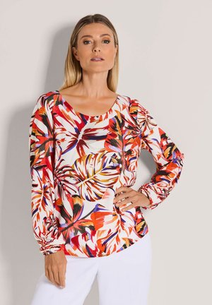 Vrouw draagt een blouse met lange mouwen met gedurfde rode, oranje, zwarte en witte tropische bladvormen, gecombineerd met een witte broek, poseert met hand op de heup.