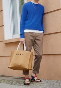 Pull bleu à côtes, t-shirt blanc, pantalon beige, sandales colorées et un grand sac en toile Marni tissé en beige avec un lettrage logo noir.