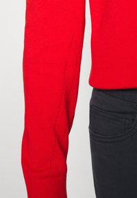 Manica di un maglione rosso lavorato a maglia con polsini a coste e cuciture decorative. Abbinato a pantaloni scuri con tasca laterale e una texture liscia.