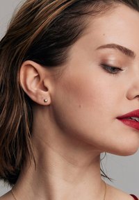 Złoty, serduszko kształtne kolczyk w uchu modelki; krótkie włosy z pasemkami; gładka cera; intensywnie czerwone usta; neutralne tło.