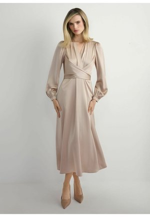 Kobieta o blond włosach, ubrana w beżową satynową sukienkę midi z długimi bufkowymi rękawami i beżowe szpilki w szpic, stojąca na jednolitym tle.
