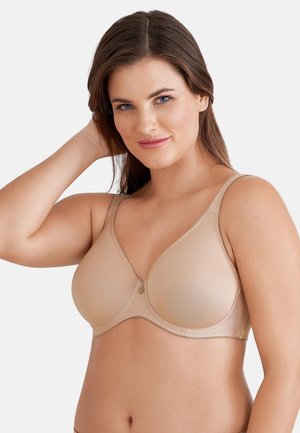 Kvinde iført en beige bh med bøjle, der smiler og rører ved sit hår mod en ensartet lys baggrund.