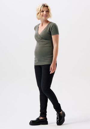 Grünes, kurzärmliges Umstandstop mit V-Ausschnitt und Raffung an den Seiten, kombiniert mit schwarzen Skinny-Jeans und Plateauschuhen.