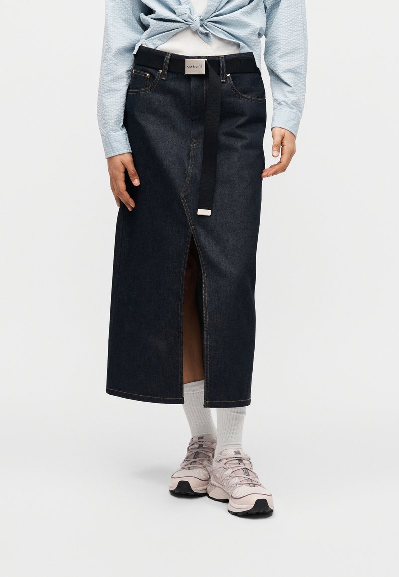 Jupe midi en denim foncé avec une fente avant, équipée d'une ceinture noire, coutures apparentes, portée avec des baskets rose clair et des chaussettes blanches.