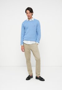 Világoskék cable-knit pulóver fehér kockás ing felett, bézs nadrág és fekete loaferek bojt részlettel. Egyszerű és klasszikus dizájn.