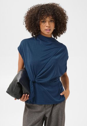 Femme aux cheveux bouclés portant un haut bleu marine drapé noué à la taille, un pantalon gris et tenant une grande pochette en cuir noir.