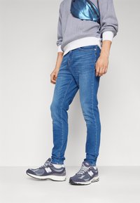 Jean en denim bleu clair avec une coupe slim, portés avec un pull gris et un sac bandoulière bleu marine, associés à des baskets grises et blanches.