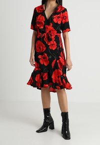 Černé zavinovací šaty zdobené velkým červeným květinovým potiskem, splývavý lem, krátké rukávy a vázání v pase. Nosit s lesklými černými kotníkovými botami.