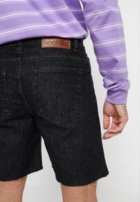 Čierne denimmové šortky s rozstrapkanými okrajmi, dvoma zadnými vreckami a hnedou koženou nášivkou značky. Nosené s fialovým prúžkovaným dlhým rukávom.