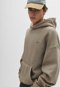 Hanorac cu glugă dintr-un amestec de bumbac de culoare taupe, cu o buzunar frontal și un detaliu subtil al logo-ului. Poartă o șapcă de culoare albastru deschis cu o margine bej.