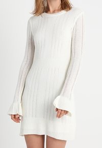 Abito bianco lavorato a maglia con maniche lunghe con cappucci svasati, coste verticali e motivi testurizzati sul corpo e sulle maniche.