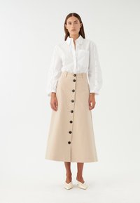 Camicia bianca con maniche a sbuffo e una gonna midi beige con bottoni neri lungo il davanti. Tessuto morbido, silhouette classica.