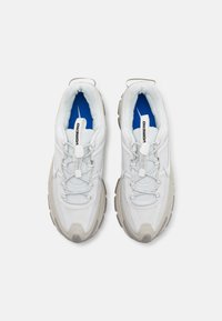 Par av vita träningsskor med tjocka grå sulor, rep-liknande snören med snabblås, och blå innersulor, sedd uppifrån på en vit bakgrund.