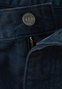 Gros plan sur un tissu denim bleu foncé avec un bouton en métal marqué « MNG DENIM » et une fermeture éclair en laiton partiellement visible.