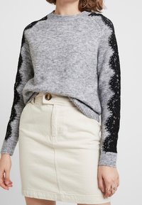 Donna che indossa un maglione grigio con dettagli di pizzo nero sulle maniche e una gonna crema a vita alta con chiusura a bottoni.