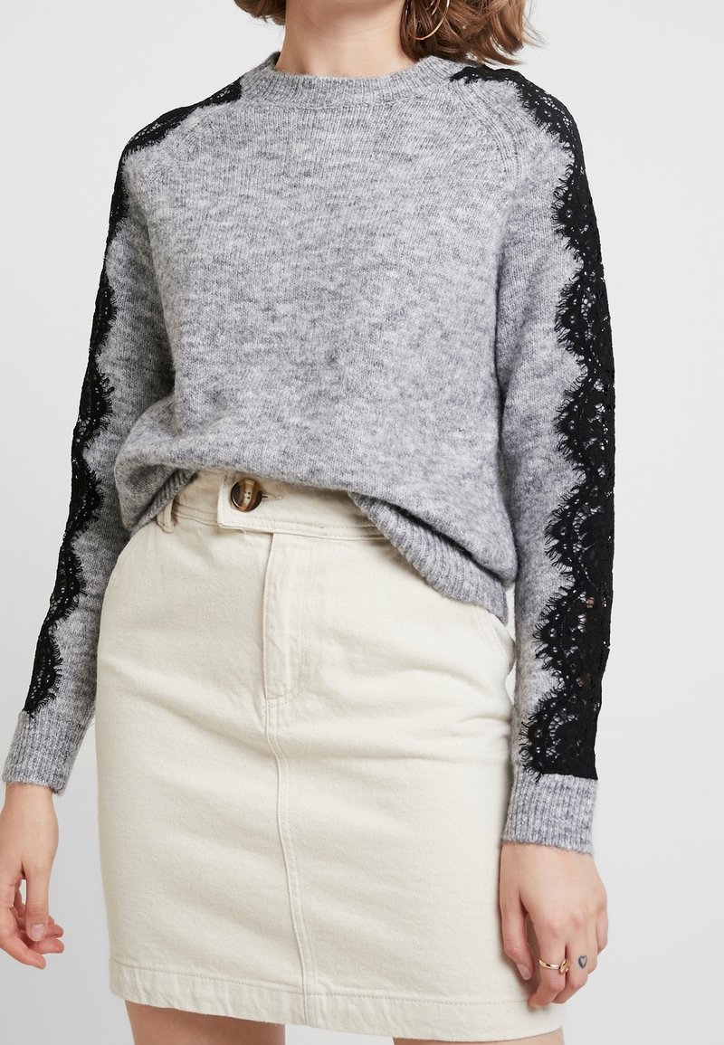 Donna che indossa un maglione grigio con dettagli di pizzo nero sulle maniche e una gonna crema a vita alta con chiusura a bottoni.