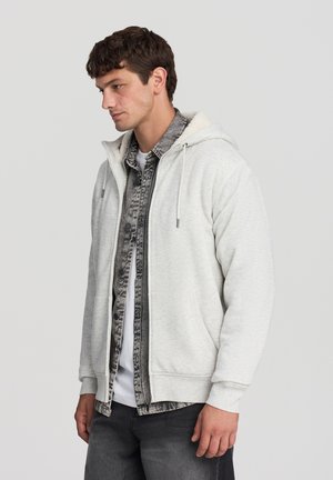 Felpa zip grigia chiara con cappuccio foderato morbido, caratterizzata da un design bicolore sopra una camicia scozzese scura e jeans grigi scuri.