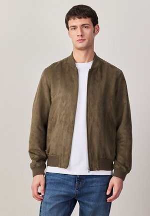 Muž nosící hnědou bomber bundu přes bílé tričko a modré džíny, stojící před jednobarevným světlým pozadím.