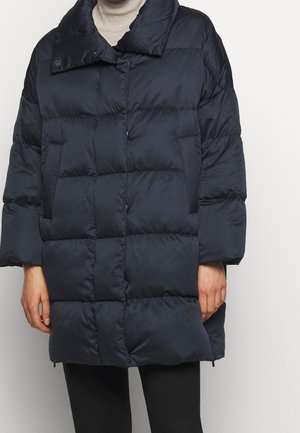 Osoba ubrana w czarną pikowaną kurtkę puchową zapinaną na napy, na beżowym golfie i czarnych spodniach, stojąca na jednolitym tle.