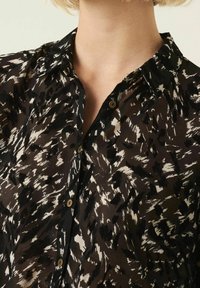 Blusa a maniche lunghe in bianco e nero con un colletto, chiusura con bottoni e tessuto trasparente; presenta un mix di forme e textures astratte.