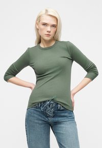 Νέα γυναίκα με ξανθά μαλλιά, που φοράει πράσινο ριμπ πλεκτό μακρυμάνικο μπλουζάκι και μπλε τζιν με κορδόνια, στέκεται με τα χέρια στη μέση μπροστά σε λευκό φόντο.