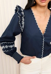 Navy blouse with ruffled, embroidered shoulders, scalloped neckline, and button closures. Striped cuffs feature floral accents.