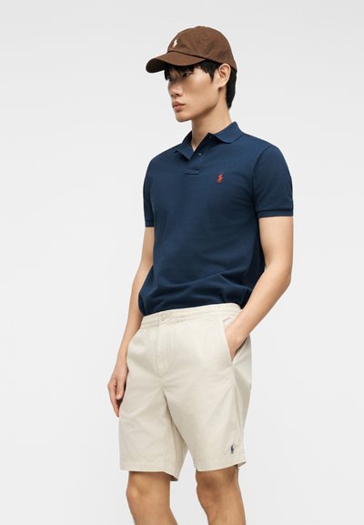 Νεαρός άνδρας με καφέ καπέλο, μπλε ναυτικό πόλο μπλουζάκι και μπεζ σορτς, στέκεται με το ένα χέρι στην τσέπη μπροστά σε απλό φόντο.