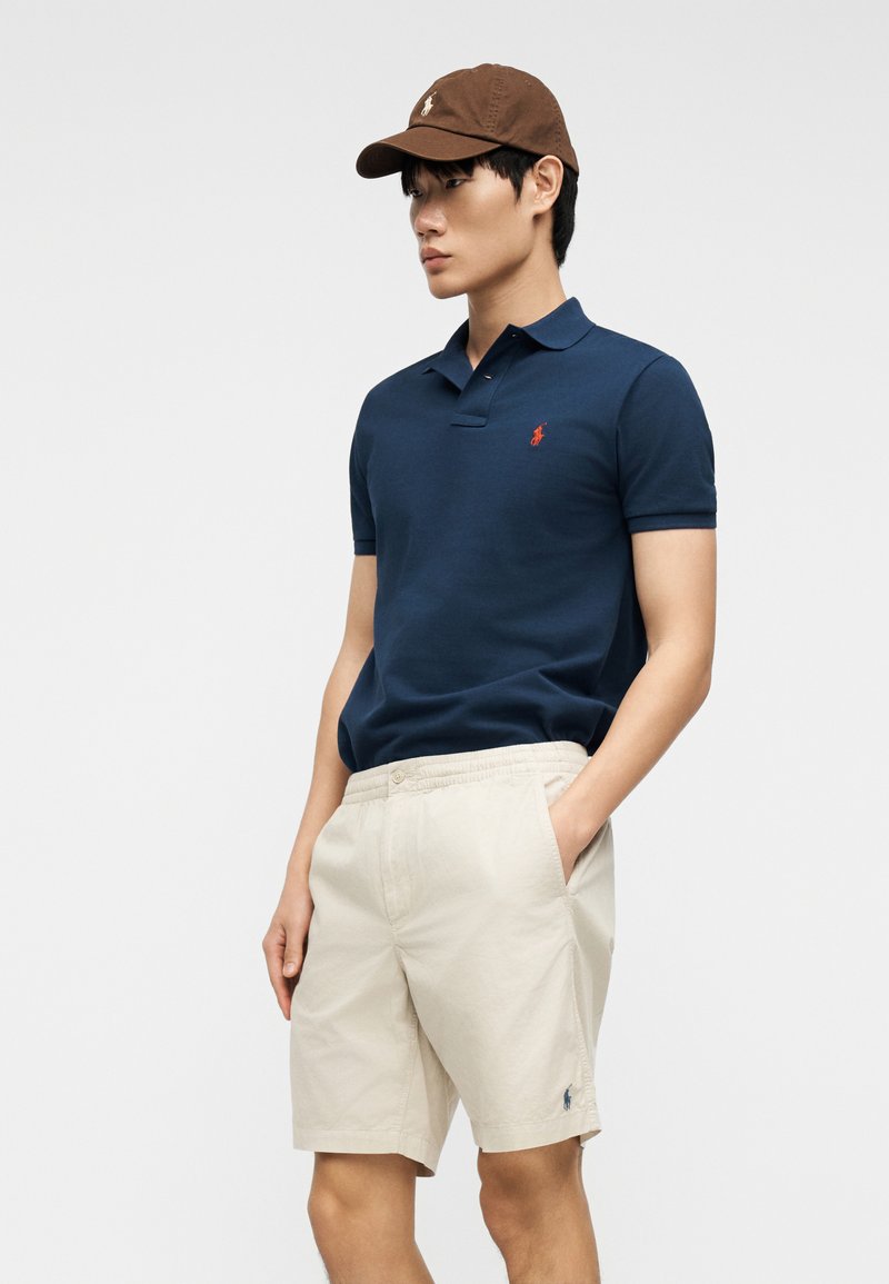 Jeune homme portant une casquette marron, un polo bleu marine et un short beige, debout avec une main dans la poche devant un fond uni.