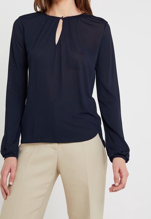 Femme portant un chemisier noir à manches longues avec une petite ouverture devant et un pantalon beige sur un fond uni.
