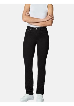 Person iført slim-fit sorte jeans, hvid ærmeløs top og hvide sneakers, stående med armene krydset mod en ensfarvet baggrund.