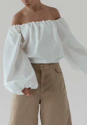 Kobieta w białej bluzce z opuszczonymi ramionami i bufiastymi rękawami oraz w beżowych spodniach z wysokim stanem i szerokimi nogawkami na jednolitym tle.