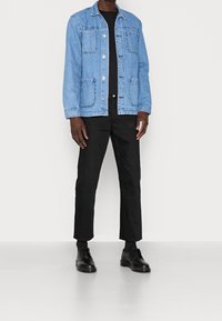 Denim jacket in light blue with buttoned front, two large pockets, and classic collar. Paired with black trousers and black shoes.