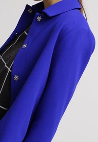 Giacca blu cobalto con design strutturato, caratterizzata da un colletto e accenti di bottoni argentati. Il tessuto testurizzato aggiunge interesse visivo.