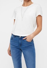 Weißes T-Shirt mit kurzen Ärmeln, kombiniert mit blauen Jeans. Das T-Shirt hat einen runden Ausschnitt; die Jeans haben eine mittlere Leibhöhe und vordere Taschen.