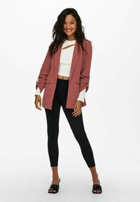 Růžový blazer s ohrnutými rukávy přes bílý cropped top a černé legíny. Oblečení doplňují černé klouzavé sandály.