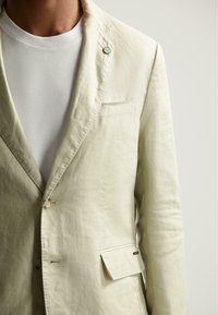 Hellbeige Leinenblazer mit einem Ein-Knopf-Verschluss, Brusttasche und einer dekorativen grünen Nadel. Glatte Textur mit lockerer Passform.